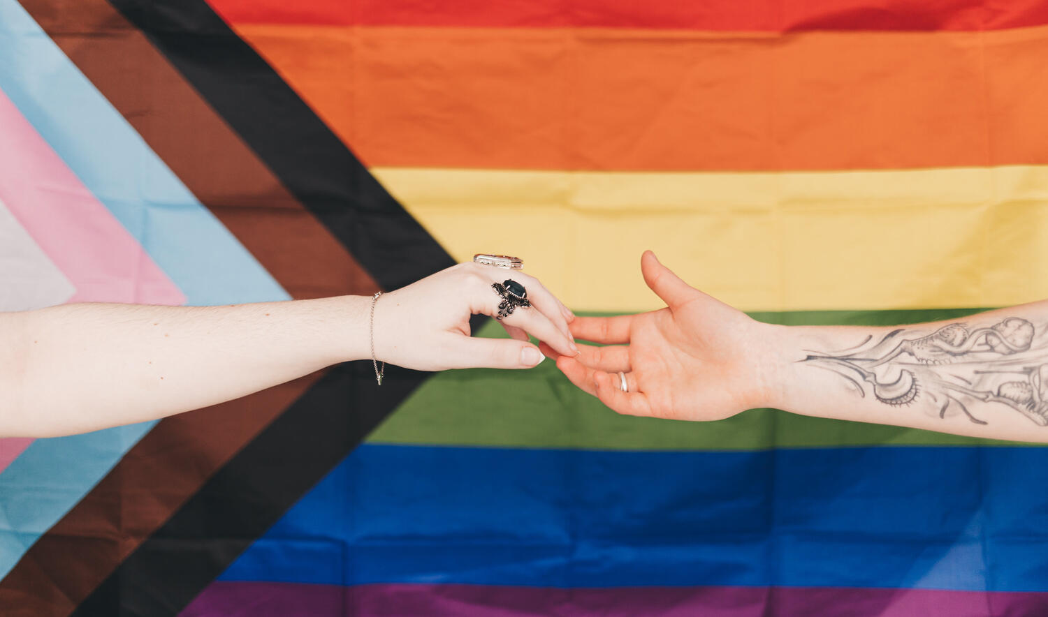 Two hands reaching for each other in front of the Progress Pride flag.